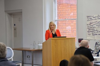 Gayle Reilly in red blazer speaking at podium